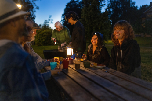 Människor sitter ute runt ett bord, i mörkret. På bordet står en elektrisk lampa. 