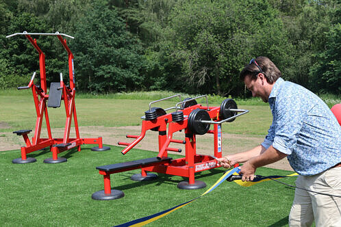 En man i blå skjorta klipper ett invigningsband med en sax. I bakgrunden två röda träningsmaskiner.